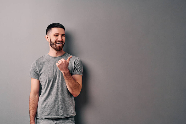 Portrait of a happy young man in gray t-shirt pointing fingers away at copy space isolated over gray background. - Фото, изображение