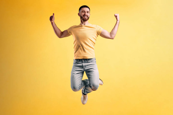 Life is beautiful! Young man dressed in a yellow t-shirt and light jeans jumping against a yellow wall shows thumbs up and smiling. - Photo, image