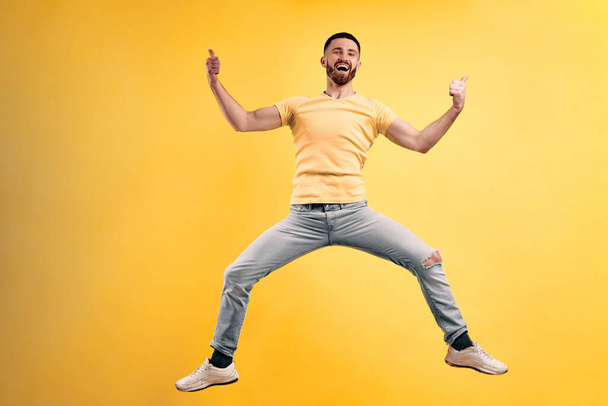Це круто! Молодий чоловік у жовтій футболці та легких джинсах стрибає на жовтому тлі стіни і тримає свої великі пальці вгору
. - Фото, зображення