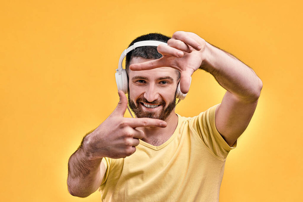 Portrait of a handsome young man in white stylish headphones showing a frame with his fingers. - Photo, Image