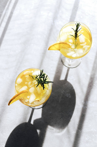 Cocktails rafraîchissants d'été avec des oranges et du romarin dans des verres en verre sur une table grise, vue sur le dessus. Boit sous un soleil éclatant, avec des ombres rudes. Soif apaisante - Photo, image