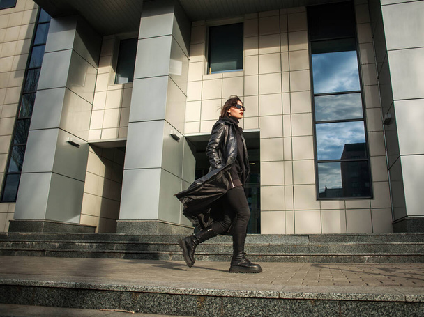 Kobieta w czarnym skórzanym płaszczu i okularach spaceruje w pośpiechu od budynku centrum biznesowego - Zdjęcie, obraz