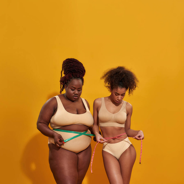 Studio shot of two african american women with different body weight measuring their waist using metric tape measure, standing together isolated over orange background - 写真・画像