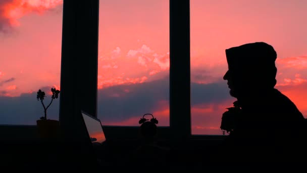Online education. Silhouette of a hacker working at a computer, a man at a table by the window at sunset, a student in a sweatshirt with a hood on his head is typing on the keyboard. Typing on a laptop - Metraje, vídeo
