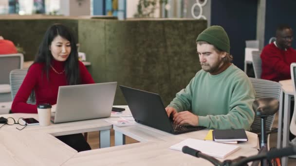 Tracking shot van een bebaarde jonge zakenman met muts en een Aziatische zakenvrouw achter een bureau in een co-working ruimte of een open kantoorruimte van een startende onderneming en werkend op laptops - Video