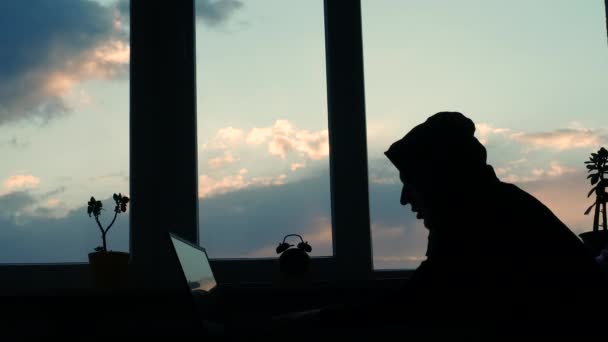 Silhouette of a hacker working at a computer, a man at a desk by the window at sunset, a student in a hoodie with a hood on his head is typing on the keyboard. Typing on a laptop. - Metraje, vídeo