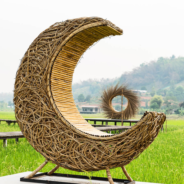 Скамейка из полулунного ротанга на зеленом рисовом поле с горным фоном и деревней. - Фото, изображение