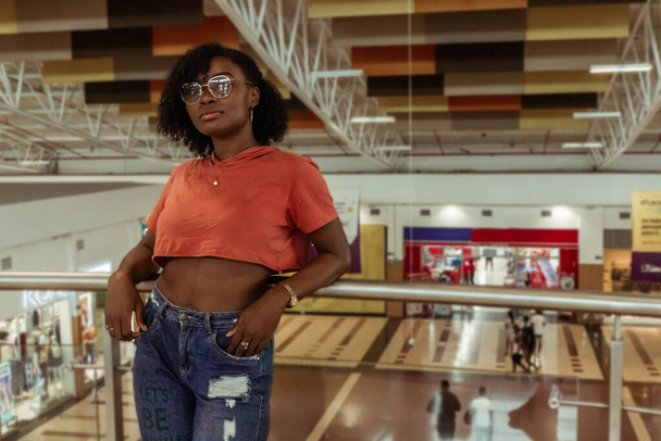 Joven mujer afro con lentes reposa en pasamanos del segundo piso del centro comercial - Foto, Imagem