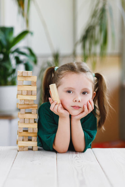 Portrait of beautiful sweet little girl in green dress with ponytails on her head holding a wooden block and wooden tower standing on a table. - Photo, image