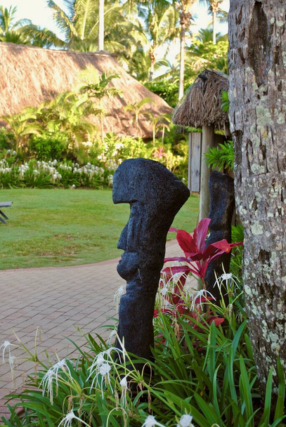 A stone tiki, white spider lilies, and a red Florica Cordyline plant a in Fiji garden. Tikis are large wooden or stone carvings or statues of human forms common throughout the Fiji islands. - Photo, Image