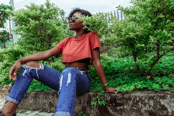 Joven bella afro con lentes de sol mirando hacia la izquierda sentada en la acera con plantas - Foto, afbeelding
