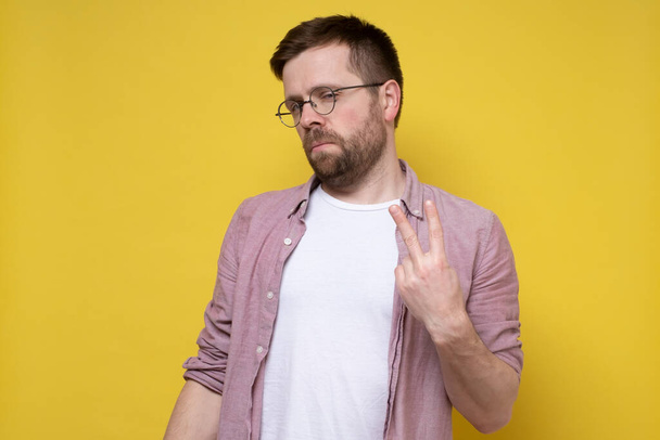 Serious man shows the number two on fingers or a victory sign and looks suspiciously at the camera. Yellow background. - Photo, Image