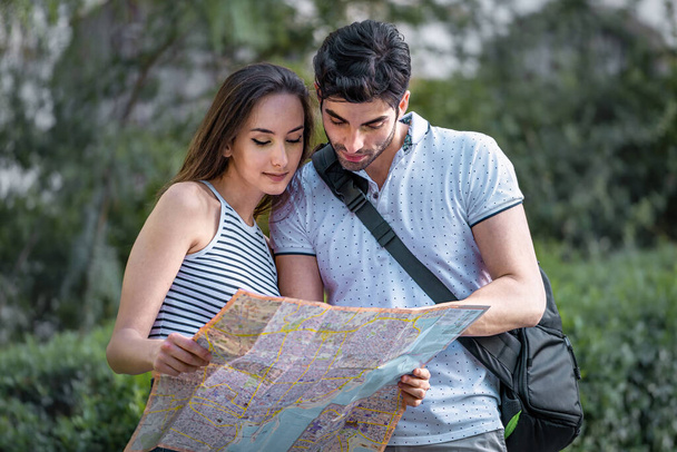 Young couple of tourists searching for a place in the map while theyre walking in the park. Travellers concept - Foto, Imagen