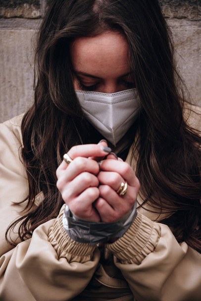Covid-19 'a karşı koruyucu bir yüz maskesi takan genç bir kadın elleri yapışkan bantla bağlanmış ve kavramsal bir görüntüyle başını eğmiş bir şekilde oturuyor. - Fotoğraf, Görsel