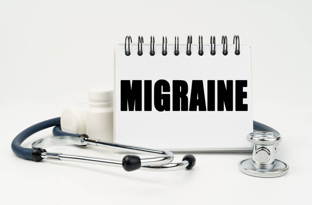 Medicine and health concept. On a white table lies a stethoscope, pills and a notebook with the inscription - MIGRAINE - Фото, изображение