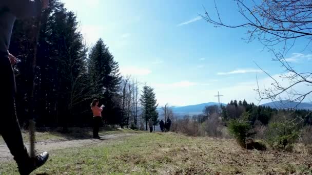 Limanowa, Poland: Rear side of a man with backpack, water bottle and a wooden stick while hiking or trekking in the polish mountains towards a landmark of metallic cross during day time - Footage, Video