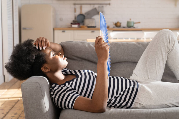 Перегріта африканка лежить на дивані використовує паперовий вентилятор для охолодження повітря страждає від гарячої температури всередині
 - Фото, зображення