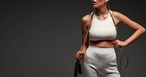 Close up view portrait of young woman with fit body holding skipping rope around her neck. Isolated grey background. Copy space. Web banner. - Photo, Image