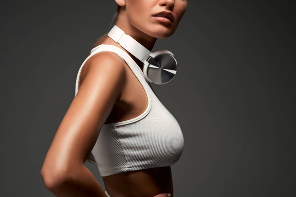 Close up view of a young pretty sporty woman posing over grey background in white athletic clothes, wearing headphones on her neck. Cropped photo. - Фото, изображение
