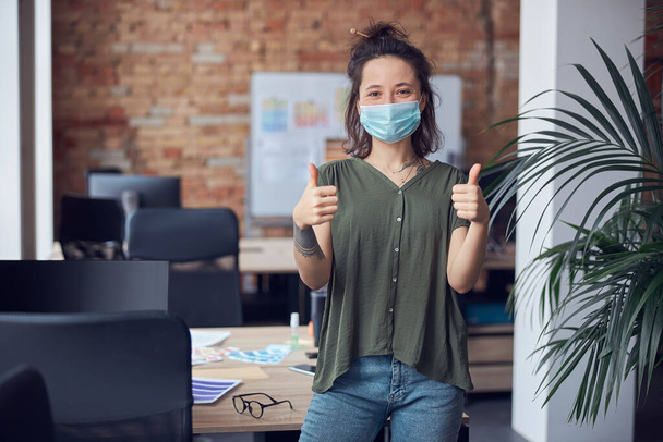 Веселая креативная женщина в защитной маске улыбается в камеру, показывает гром, стоя в современном зеленом офисе в дневное время - Фото, изображение
