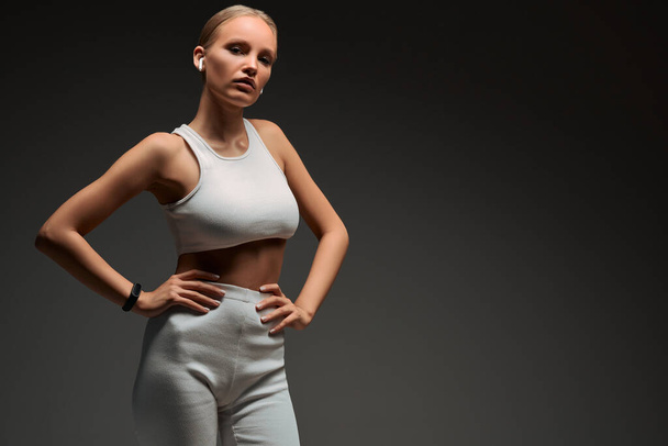 Sports blonde girl in white training clothes with headphones in the studio on a grey background. The concept of training, healthy lifestyle, portrait of a fitness instructor. Copy space. - Zdjęcie, obraz