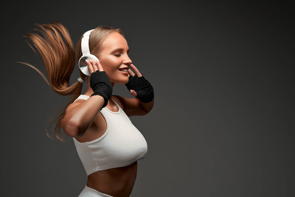 Woman with perfect abs wearing headphones and white clothes standing in gym isolated grey background. Copy space. - Foto, Imagem