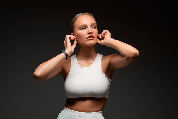 Training with music. Portrait of young fitness woman wearing wireless earbuds while standing isolated over grey background, concept of sport, fitness and healthy lifestyle. - Foto, imagen