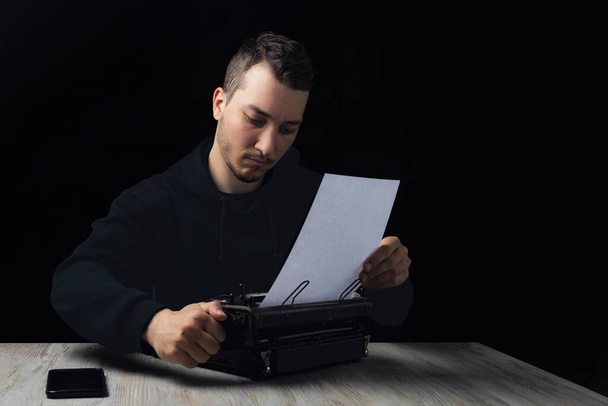 Young handsome man in black hoodie inserts paper into typewriter on black background in low key - Photo, Image