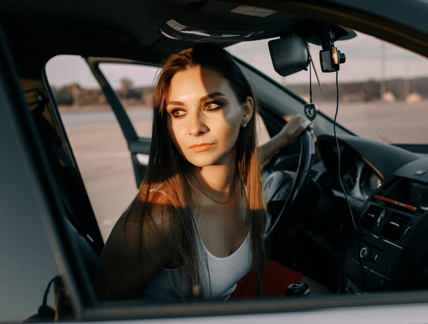 Beautiful young girl driving a car in the evening in the sunset sun on an empty parking lot - 写真・画像