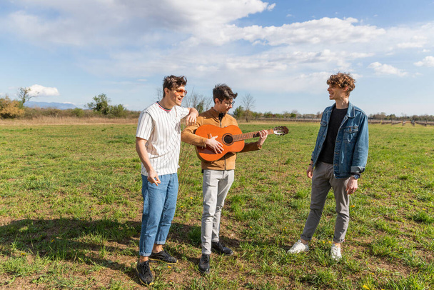 kolme ystävää kentällä kokoontuvat laulamaan ja soittamaan kitaraa iloisesti yhdessä. - Valokuva, kuva