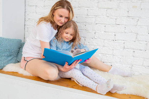 Happy caucasian mother with her daughter reading a book and smiling while sitting on the floor in the living room - Zdjęcie, obraz