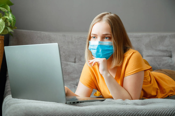 Woman in medical protective mask work using laptop at Home office lies on couch. Teen girl in mask Online learning education via laptop covid 19 lockdown time. Remote work in Coronavirus pandemic. - Foto, imagen