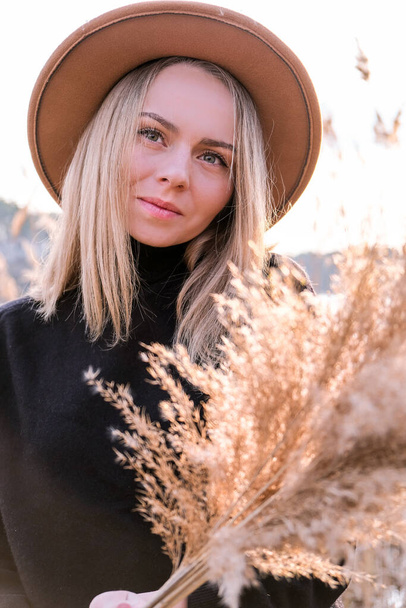 European blonde woman with beige hat in black sweater in the countryside. Golden hour, cottagecore. Local travel. Slow living. Mental health. Pampas grass - Zdjęcie, obraz