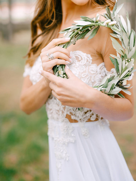 白い刺繍のドレスの美しい花嫁は横に立って、彼女の手に緑のオリーブの枝の花輪を保持しています - 写真・画像