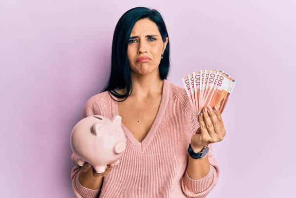 Young caucasian woman holding norwegian krona banknotes and piggy bank depressed and worry for distress, crying angry and afraid. sad expression.  - Foto, Imagen