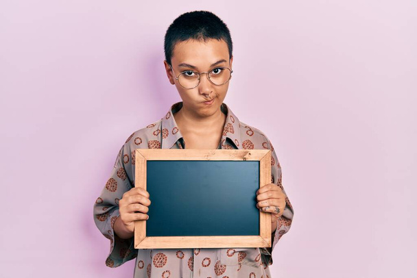 Beautiful hispanic woman with short hair holding blackboard skeptic and nervous, frowning upset because of problem. negative person.  - Foto, Imagen