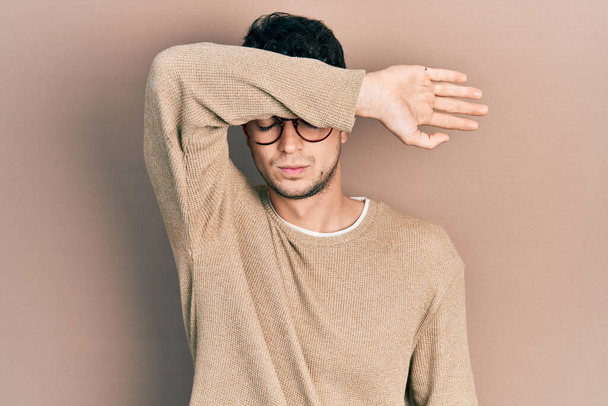 Jonge Spaanse man draagt nonchalante kleren en een bril die ogen bedekt met een arm, en er ernstig en verdrietig uitziet. gezichtsloosheid, verstoppertje en afwijzing  - Foto, afbeelding