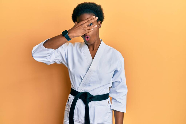 Jovem afro-americana vestindo kimono de karatê e faixa preta espreitando em choque cobrindo rosto e olhos com a mão, olhando através de dedos com expressão embaraçosa.  - Foto, Imagem