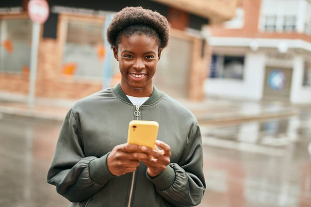 Jeune fille afro-américaine souriant heureux en utilisant un smartphone à la ville - Photo, image