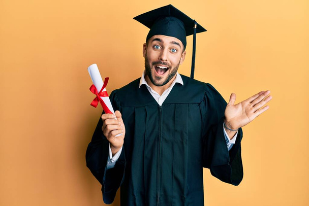 Mezuniyet cübbesi giyen genç İspanyol adam diplomasını elinde tutarak başarıyı mutlu bir gülümsemeyle ve kazanan yüz ifadesiyle kutluyor.  - Fotoğraf, Görsel