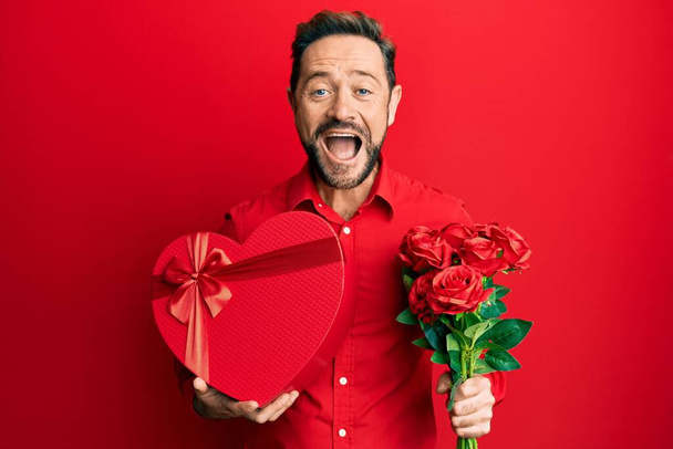 Middle age man holding anniversary present and bouquet of flowers smiling and laughing hard out loud because funny crazy joke.  - Photo, Image