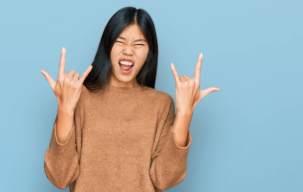 Hermosa mujer asiática joven usando suéter de invierno casual gritando con expresión loca haciendo símbolo de roca con las manos arriba. Estrella de la música. concepto pesado.  - Foto, Imagen
