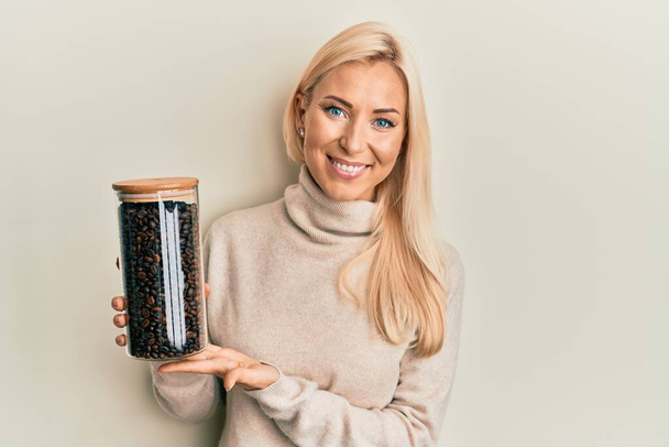 Jeune femme blonde tenant un pot avec des grains de café regardant positif et heureux debout et souriant avec un sourire confiant montrant des dents  - Photo, image