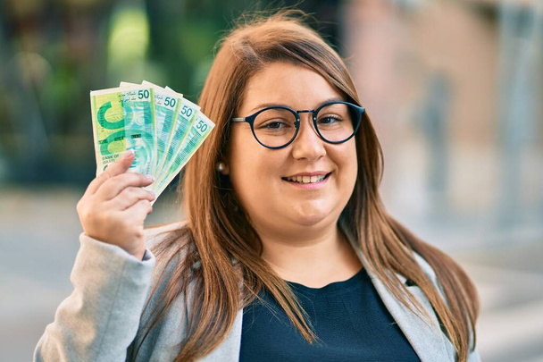 Młoda Hiszpanka plus bizneswoman wielkości uśmiechnięta szczęśliwa trzymając w ręku banknoty Israel Shekels w mieście. - Zdjęcie, obraz