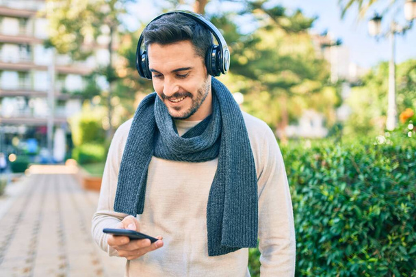 Jeune homme hispanique souriant heureux en utilisant smartphone et écouteurs au parc. - Photo, image