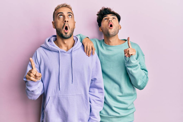 Homosexual gay couple standing together wearing casual clothes amazed and surprised looking up and pointing with fingers and raised arms.  - 写真・画像