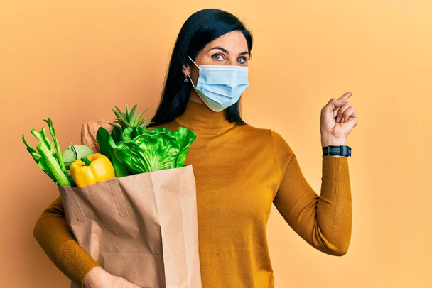 Young caucasian woman wearing medical mask holding groceries paper bag smiling happy pointing with hand and finger to the side  - Foto, Imagen
