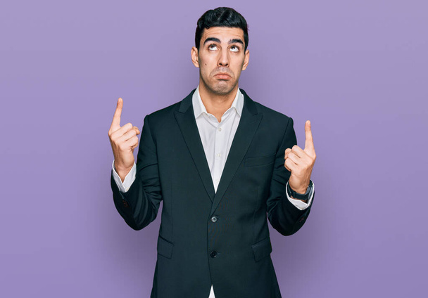 Schöner hispanischer Mann in Businesskleidung, der traurig und aufgebracht nach oben zeigt, mit den Fingern die Richtung anzeigt, unglücklich und deprimiert.  - Foto, Bild