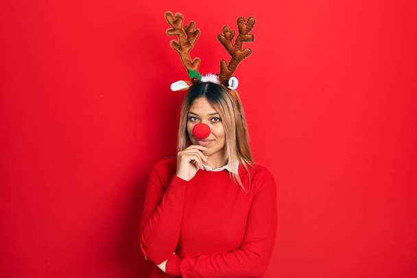 Όμορφη Ισπανίδα γυναίκα φορώντας ελάφι χριστουγεννιάτικο καπέλο και κόκκινη μύτη που αναζητούν αυτοπεποίθηση στην κάμερα με χαμόγελο με σταυρωμένα χέρια και το χέρι έθεσε στο πηγούνι. θετική σκέψη.  - Φωτογραφία, εικόνα