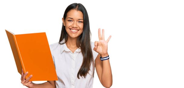 Beautiful hispanic woman reading book doing ok sign with fingers, smiling friendly gesturing excellent symbol  - Foto, Bild